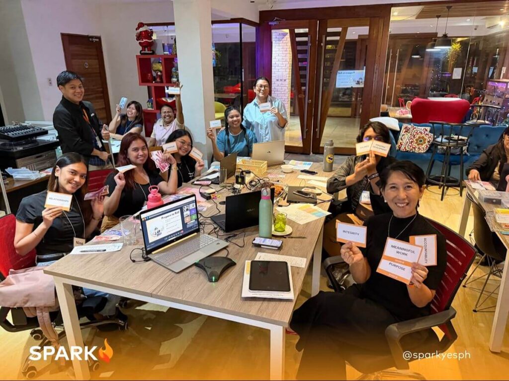 Educators participate in a collaborative learning workshop while holding activity cards around a table with laptops and training materials