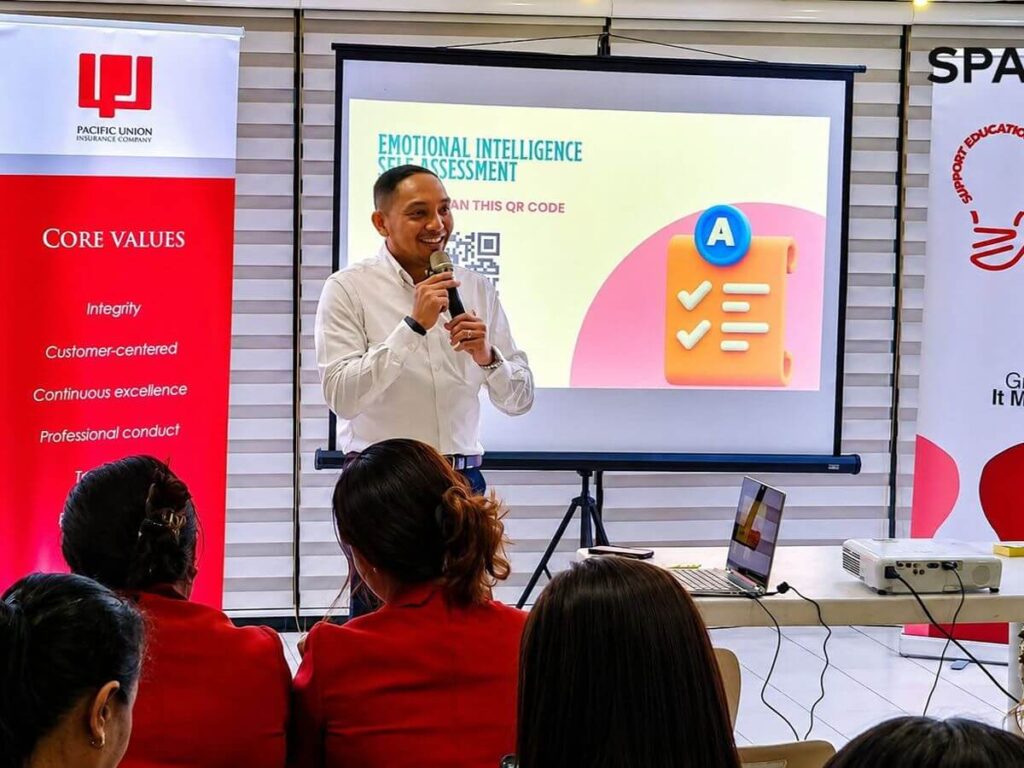 A facilitator leads an emotional intelligence self-assessment session for educators using a microphone and presentation screen